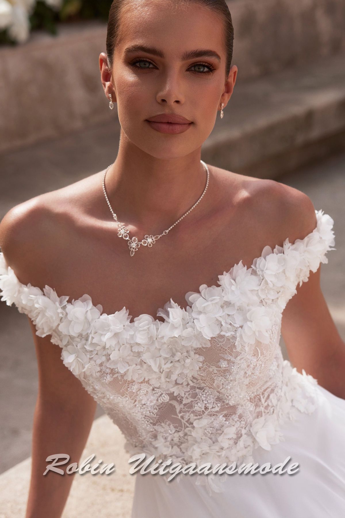 Close-up van de Off-shoulder trouwjurk met kanten lijfje,  een bloemen versierde halslijn, sluikvallende chiffon rok met semi lange sleep
