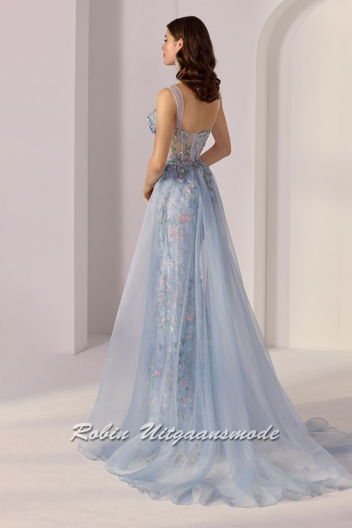 Achterkant van de Lichtblauwe galajurk volledig versierd met geborduurde bloemen, pailletten, een korset lijfje met diep uitgesneden bustelijn, lange rok met hoge split en een organza overrok